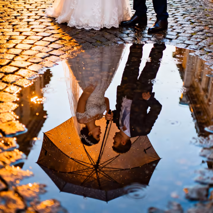 A stunning wedding photograph of a couple reflected in a puddle, holding an umbrella as rain falls around them, creating a romantic and artistic scene.